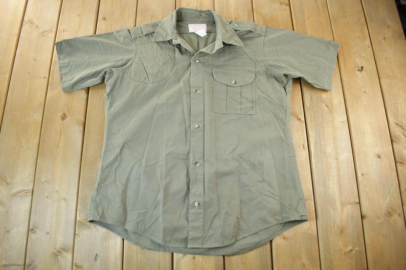 Peut inclure: Chemise vert olive &agrave; manches courtes avec deux poches de poitrine et une fermeture boutonn&eacute;e. La chemise a un col et des &eacute;paulettes sur les &eacute;paules. Le tissu semble &ecirc;tre l&eacute;ger.