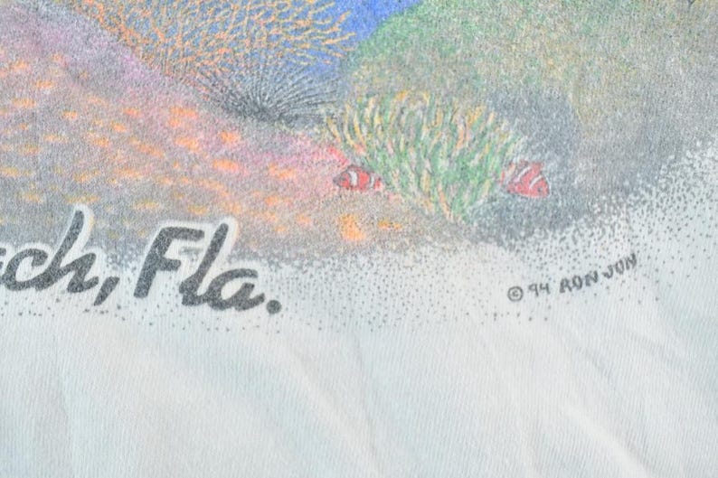 Puede incluir: Camiseta blanca con una colorida escena submarina con corales, peces y el texto "Beach, Fla.". El dise&ntilde;o incluye la marca de derechos de autor y "94 RON JON" en negro. La obra de arte representa un vibrante arrecife de coral.