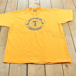 Peut inclure: Un t-shirt jaune vif à manches courtes avec une impression graphique bleue sur la poitrine. Le motif représente le contour d'un globe avec la lettre T au centre, entouré d'une bannière portant le texte bleu Toastmasters International.
