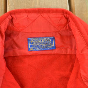 May include: Close-up of a red wool jacket collar with a blue and white label. The label reads "Pendleton Woolen Mills" and includes text about the fabric content and origin. The jacket is made in the U.S.A.