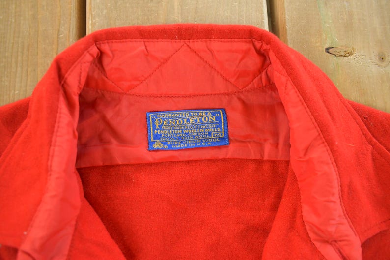 May include: Close-up of a red wool jacket collar with a blue and white label. The label reads "Pendleton Woolen Mills" and includes text about the fabric content and origin. The jacket is made in the U.S.A.