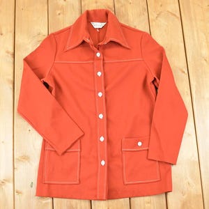 Op de afbeelding: Een vintage oranje jasje met een kraag, lange mouwen en witte stiksels. De jas heeft twee voorzakken en witte knopen aan de voorkant. Het jasje wordt gepresenteerd op een houten oppervlak.