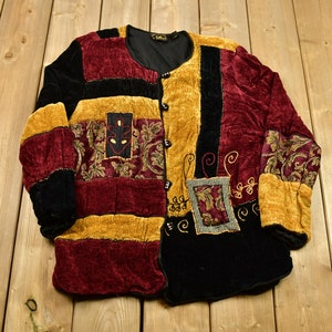 Op de afbeelding: Een patchwork jas van bordeaux, goud en zwart fluweel. De jas heeft een ronde kraag, een knoopsluiting en decoratieve geborduurde patches. De mouwen en voorpanelen hebben een mix van texturen en patronen, wat een unieke vintage look creëert.