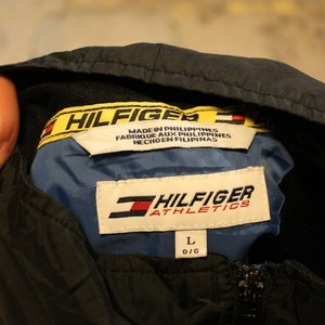 Pu&ograve; includere: Una giacca nera con cappuccio con un'etichetta Hilfiger Athletics gialla e nera. L'etichetta dice "Hilfiger Athletics" e "Made in Philippines, Fabrique aux Philippines, Hecho en Filipinas". La giacca ha un'etichetta di taglia che dice "L G/G".