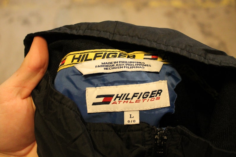Pu&ograve; includere: Una giacca nera con cappuccio con un'etichetta Hilfiger Athletics gialla e nera. L'etichetta dice "Hilfiger Athletics" e "Made in Philippines, Fabrique aux Philippines, Hecho en Filipinas". La giacca ha un'etichetta di taglia che dice "L G/G".