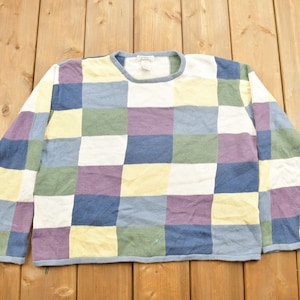 May include: A colorful patchwork sweater with a crew neck and long sleeves. The sweater is made of a soft, knitted fabric and features a checkered pattern in shades of blue, green, purple, and yellow.