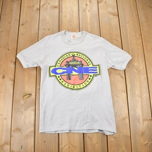 May include: A light gray t-shirt with a graphic of a yellow, red, and blue logo that says "Canadian National Exhibition" with a stylized archway in the center.