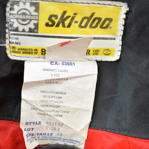 May include: A yellow and black Ski-Doo label with the text "ski-doo" in large white letters. The label also includes the text "Bombardier" and "Marques de Trade Marks of B". Below the label is a white tag with the text "CA: 03661", "Femmes/Ladies", "2 PCE", "Habit/Suit", "Shell/Exterieur", "100% Nylon", "Sinine Doublure", "100% Nylow", "Entre noublora/Inter Linine", "100% Polyester", "Style 491157", "Lot 1-063", "Size/Taille 12", and "Exclusive of Trim/Garniture Made in Canada/Fabrique au Canada".