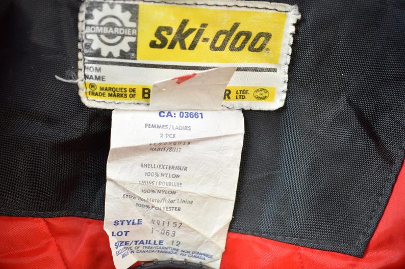 May include: A yellow and black Ski-Doo label with the text "ski-doo" in large white letters. The label also includes the text "Bombardier" and "Marques de Trade Marks of B". Below the label is a white tag with the text "CA: 03661", "Femmes/Ladies", "2 PCE", "Habit/Suit", "Shell/Exterieur", "100% Nylon", "Sinine Doublure", "100% Nylow", "Entre noublora/Inter Linine", "100% Polyester", "Style 491157", "Lot 1-063", "Size/Taille 12", and "Exclusive of Trim/Garniture Made in Canada/Fabrique au Canada".