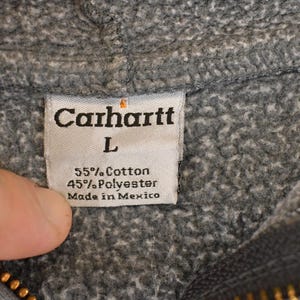 Puede incluir: Primer plano de una etiqueta de sudadera con capucha Carhartt gris. La etiqueta dice "Carhartt" encima de la talla "L". Tambi&eacute;n indica la composici&oacute;n del tejido: 55% algod&oacute;n, 45% poli&eacute;ster y "Made in Mexico".