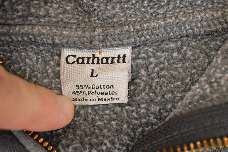 Puede incluir: Primer plano de una etiqueta de sudadera con capucha Carhartt gris. La etiqueta dice "Carhartt" encima de la talla "L". Tambi&eacute;n indica la composici&oacute;n del tejido: 55% algod&oacute;n, 45% poli&eacute;ster y "Made in Mexico".