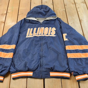 May include: A navy blue hooded jacket with the word "ILLINOIS" in orange block letters across the chest. The jacket has orange stripes on the sleeves and a matching orange and white striped hem. The jacket is zipped up.