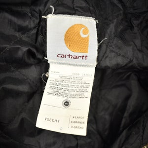 Może przedstawiać: Czarna kurtka Carhartt z białą metką z napisem "Carhartt" i logo Carhartt. Metka zawiera r&oacute;wnież rozmiar "X-Large" i numer stylu "0699 362611".
