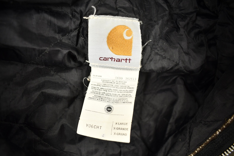 Może przedstawiać: Czarna kurtka Carhartt z białą metką z napisem "Carhartt" i logo Carhartt. Metka zawiera r&oacute;wnież rozmiar "X-Large" i numer stylu "0699 362611".