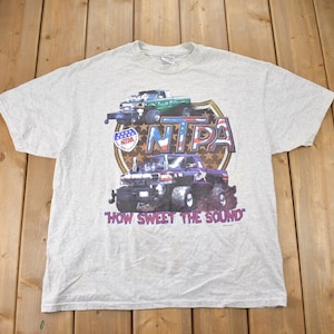 以下が含まれることがあります： 2台のモンスタートラックと「NITPA」と「HOW SWEET THE SOUND」の文字が描かれたグレーのTシャツです。