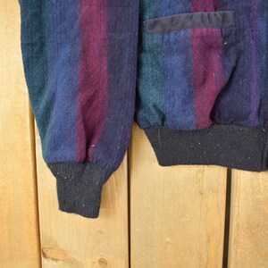 Puede incluir: Primer plano de un c&aacute;rdigan vintage con un dise&ntilde;o a rayas en tonos azules, morados y verdes. El c&aacute;rdigan tiene un dobladillo y pu&ntilde;os de canal&eacute; negros, con botones negros y un bolsillo. La tela parece ser una mezcla de lana.