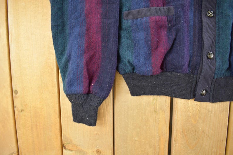 Puede incluir: Primer plano de un c&aacute;rdigan vintage con un dise&ntilde;o a rayas en tonos azules, morados y verdes. El c&aacute;rdigan tiene un dobladillo y pu&ntilde;os de canal&eacute; negros, con botones negros y un bolsillo. La tela parece ser una mezcla de lana.