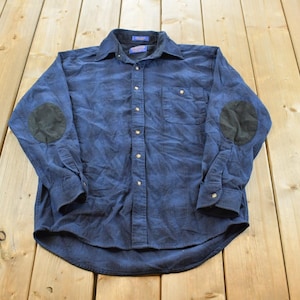 May include: A long-sleeve button-down shirt in a dark blue shade with a subtle plaid pattern. The shirt features a pointed collar, a single chest pocket on the left side, and contrasting dark circular elbow patches on both sleeves.