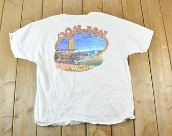 May include: White t-shirt featuring a colorful graphic of a vintage woody station wagon, a surfboard, and the text "Ron Jon" above the car. The graphic is set against a blue sky and ocean backdrop, with the text "Since 1963" below.