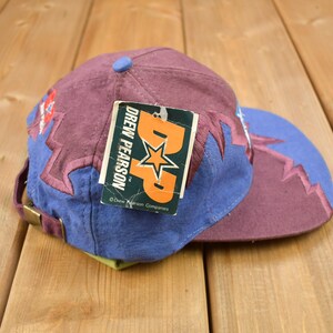 May include: A purple and blue baseball cap with a lightning bolt design. The cap has a tag attached that reads "Drew Pearson TM Drew Pearson Companies".