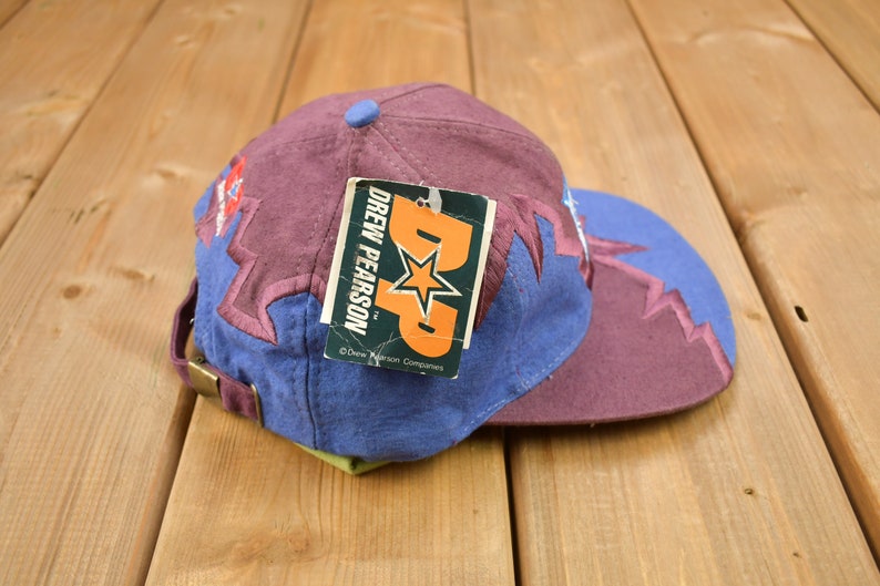 May include: A purple and blue baseball cap with a lightning bolt design. The cap has a tag attached that reads "Drew Pearson TM Drew Pearson Companies".