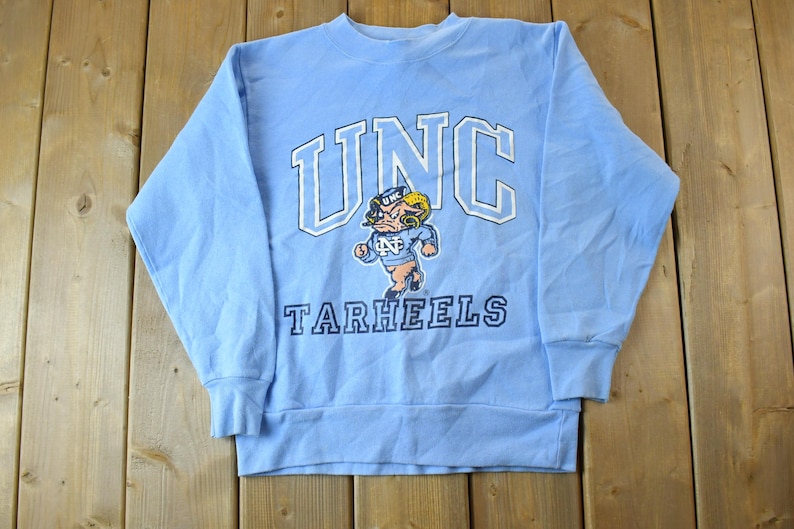 May include: A light blue sweatshirt with the text "UNC Tarheels" and a cartoon mascot in the center.