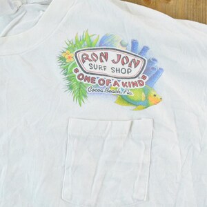 Puede incluir: Camiseta blanca con bolsillo, con un gr&aacute;fico colorido de un pez tropical, coral y el texto "Ron Jon Surf Shop ONE OF A KIND Cocoa Beach, Fla."