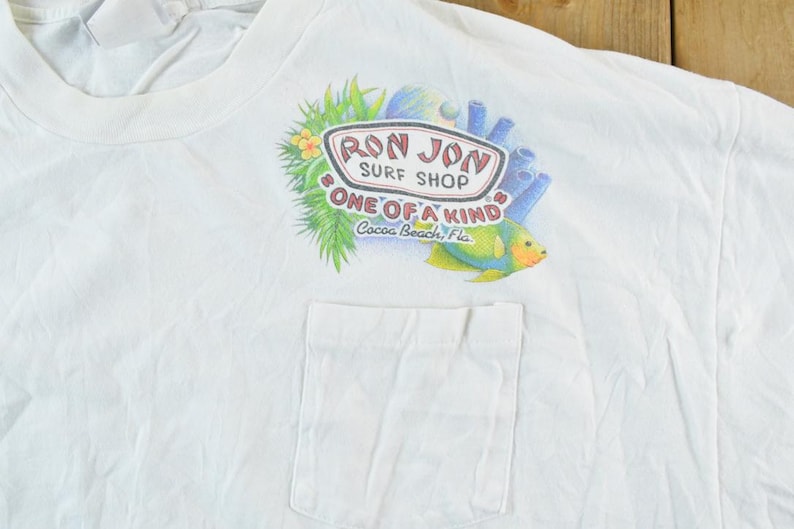 Puede incluir: Camiseta blanca con bolsillo, con un gr&aacute;fico colorido de un pez tropical, coral y el texto "Ron Jon Surf Shop ONE OF A KIND Cocoa Beach, Fla."