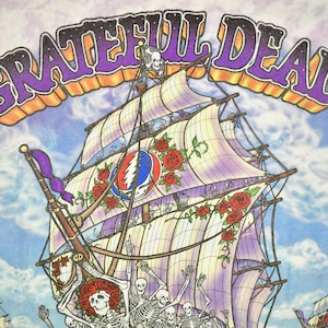 May include: A tie-dye t-shirt featuring a colourful illustration of a ship with skeletons on deck. The ship has a large sail with the Grateful Dead logo and roses. The text "GRATEFUL DEAD" is printed in purple and orange letters across the top of the image.