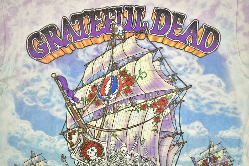 May include: A tie-dye t-shirt featuring a colourful illustration of a ship with skeletons on deck. The ship has a large sail with the Grateful Dead logo and roses. The text "GRATEFUL DEAD" is printed in purple and orange letters across the top of the image.