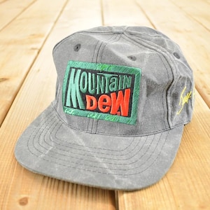 Puede incluir: Gorra de béisbol gris con visera plana. La parte delantera presenta un parche rectangular con las palabras "MOUNTAIN DEW" en verde, rojo y blanco. La gorra tiene un diseño cosido y un detalle bordado amarillo en el lateral.