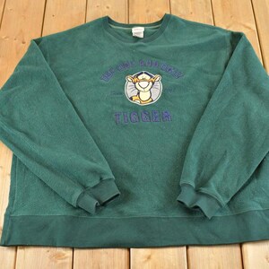 Puede incluir: Una sudadera de forro polar de manga larga en color verde oscuro con un gráfico bordado del personaje Tigger centrado en el pecho. El texto sobre el personaje dice The Only Tigger y la palabra Tigger está bordada en morado bajo el gráfico.