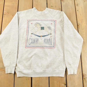 Peut inclure: Un sweat-shirt gris clair avec un col ras du cou et des manches longues. Le devant présente un motif rectangulaire avec un cheval à bascule et les mots "Country Rockin" en écriture cursive. Le motif a des accents rouges, bleus et beiges.