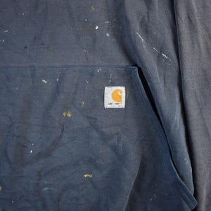K&ouml;nnte beinhalten: Ein marineblauer Kapuzenpullover mit Fronttasche und dem Carhartt-Logo-Patch. Der Stoff ist mit wei&szlig;en und gelben Farbspritzern &uuml;bers&auml;t, was ihm eine abgenutzte Arbeitskleidungs&auml;sthetik verleiht.