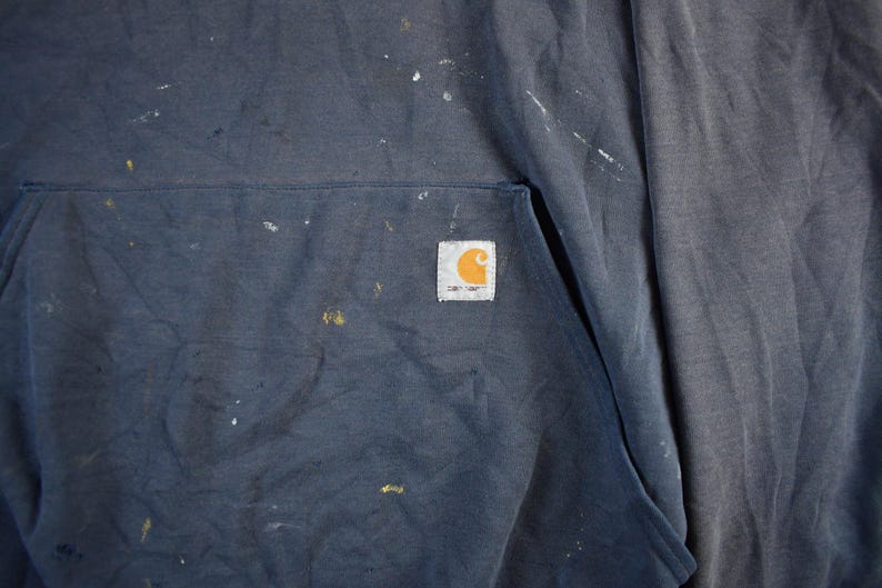 K&ouml;nnte beinhalten: Ein marineblauer Kapuzenpullover mit Fronttasche und dem Carhartt-Logo-Patch. Der Stoff ist mit wei&szlig;en und gelben Farbspritzern &uuml;bers&auml;t, was ihm eine abgenutzte Arbeitskleidungs&auml;sthetik verleiht.