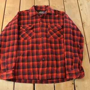 May include: A red and black checked flannel shirt with two chest pockets and a button-down front. The shirt has long sleeves and a classic collar. The fabric appears to be a medium weight, suitable for cooler weather. The shirt is laid flat on a wooden surface.