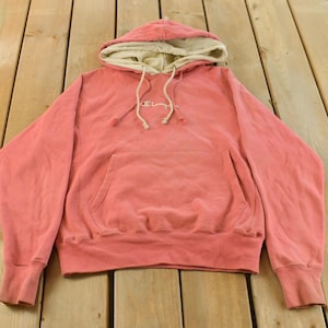 May include: A coral Champion hoodie with a cream hood lining and drawstring. The hoodie has a front pocket and the Champion logo embroidered on the chest. The garment is made of a soft, comfortable material.