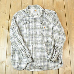 May include: A long-sleeved, button-down shirt with a plaid pattern in shades of gray, white, and blue. The shirt has two chest pockets and a collar. The Carhartt logo is visible on the chest pocket.