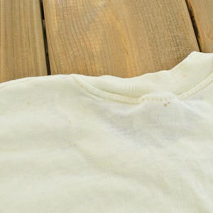 May include: A cream-colored t-shirt featuring a ribbed crew neckline and short sleeves. The fabric displays a light, off-white tone with subtle, small brown spots scattered across the shoulder and collar area, indicating signs of wear.