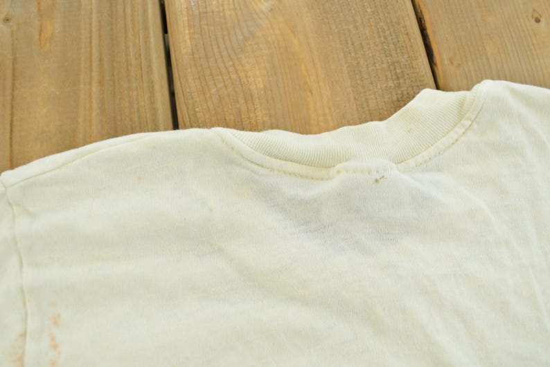 May include: A cream-colored t-shirt featuring a ribbed crew neckline and short sleeves. The fabric displays a light, off-white tone with subtle, small brown spots scattered across the shoulder and collar area, indicating signs of wear.