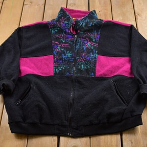 Puede incluir: Una chaqueta de forro polar negra con un patrón geométrico rosa y azul. La chaqueta tiene una cremallera y dos bolsillos.