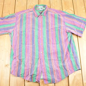 Pode incluir: Camisa de botão de manga curta com listras verticais em roxo, verde, vermelho e branco. A camisa tem dois bolsos no peito e uma gola. A camisa está estendida em uma superfície de madeira.