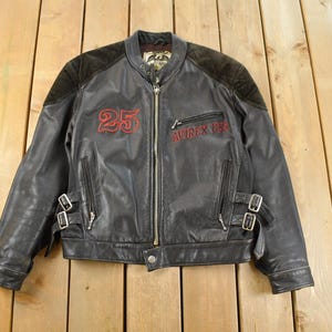 May include: Black leather jacket with a zip-up front, featuring the number "25" in red on the chest and "AVIREX USA" in red on the upper chest. The jacket has black suede shoulder patches and silver buckles.