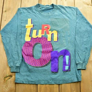 May include: Teal long-sleeve shirt with a graphic print of the word "turn on" in colorful, stylized letters.