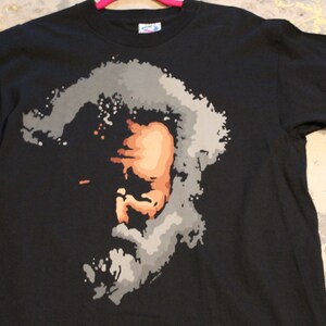 May include: Black t-shirt with a stylized portrait of a man with a beard and mustache. The portrait is made up of gray, brown, and white paint splatters.