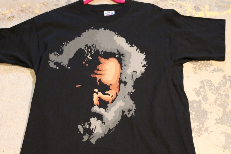May include: Black t-shirt with a stylized portrait of a man with a beard and mustache. The portrait is made up of gray, brown, and white paint splatters.