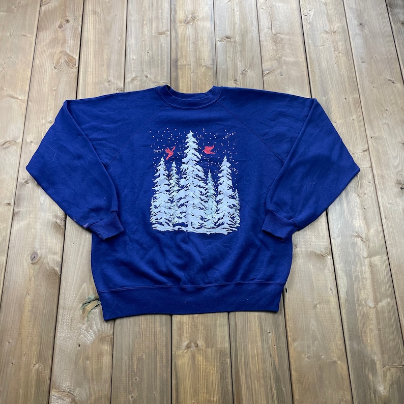 Op de afbeelding: Een marineblauw sweatshirt met een witte winterse afbeelding van een groep dennenbomen en rode kardinalen die erboven vliegen. De afbeelding is gecentreerd op het sweatshirt.
