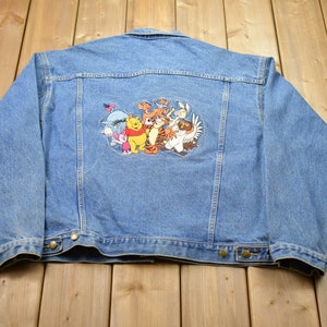 Op de afbeelding: Een blauwe denimjas met een geborduurde patch van Winnie de Poeh en zijn vrienden op de rug.