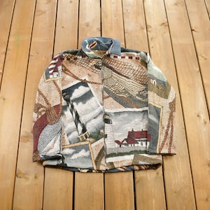May include: A brown, beige, and blue fleece jacket with a zipper closure. The jacket features a tapestry-like design with images of lighthouses, boats, and seascapes.