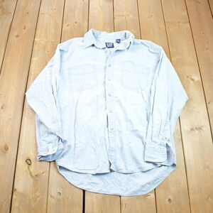 May include: A light blue long-sleeved denim shirt with a button-down collar and a single chest pocket. The shirt has the word "GAP" embroidered on the left side of the chest.